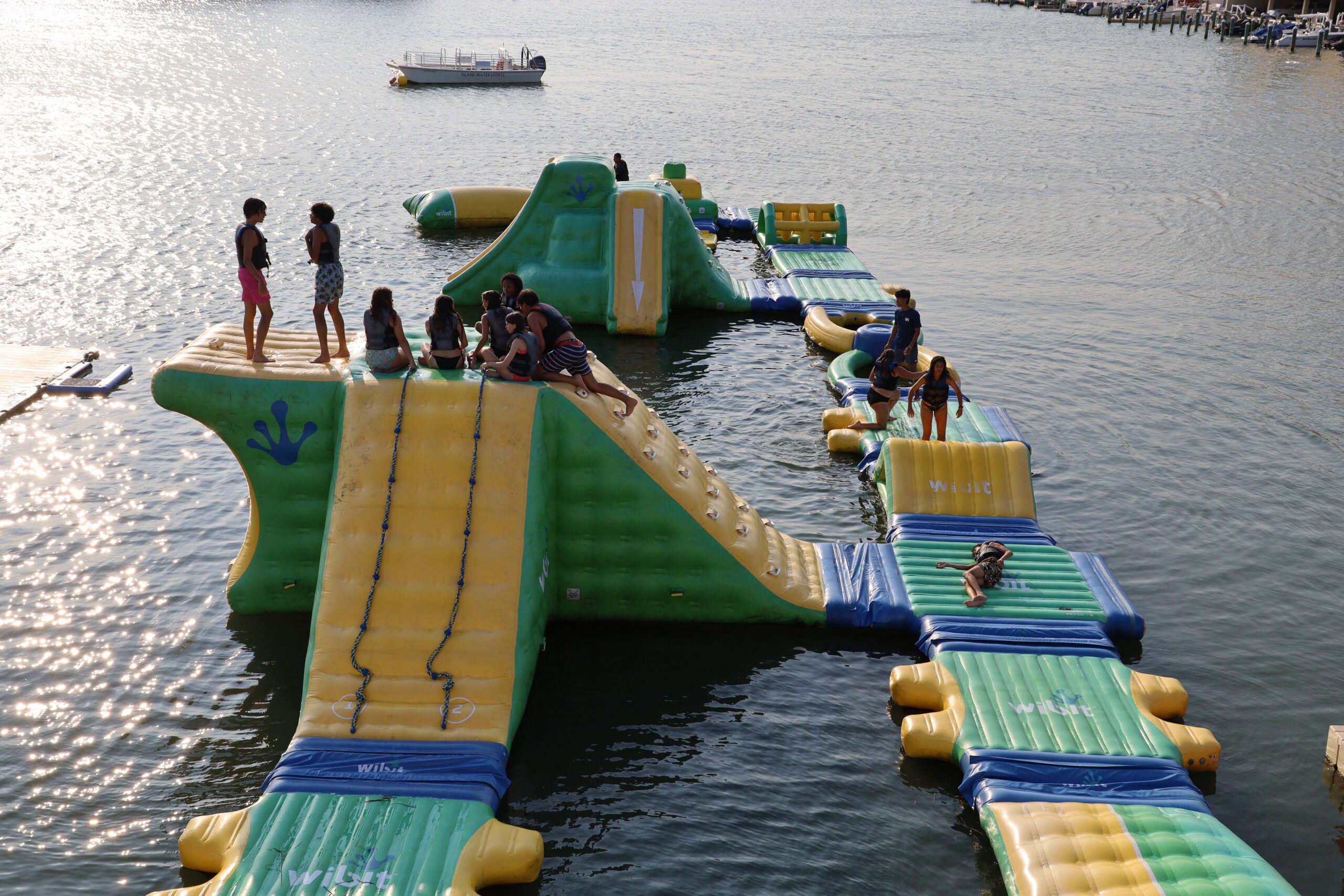 Elevated view of the full inflatable aqua park at golden hour with multiple groups of kids playing on the climbing towers, slides, and connecting platforms in Stone Harbor’s back bay