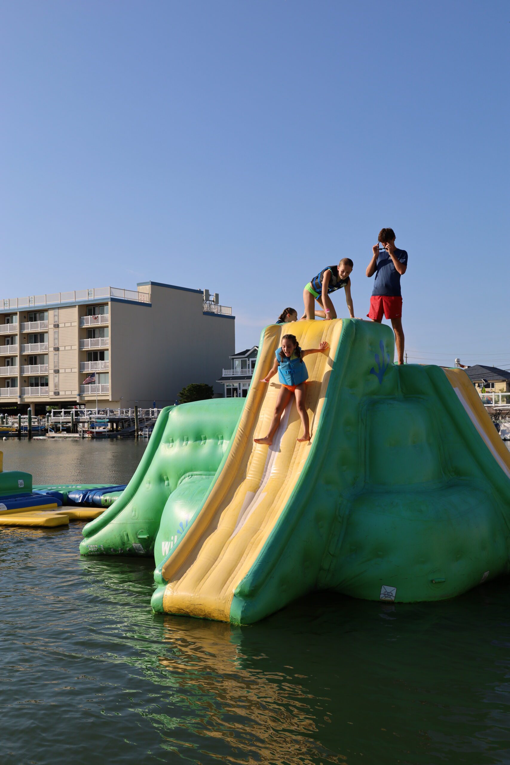 Two young guests sliding down the inflatable slide at the Stone Harbor aqua park on a clear sunny day while a teen lifeguard watches from the top
