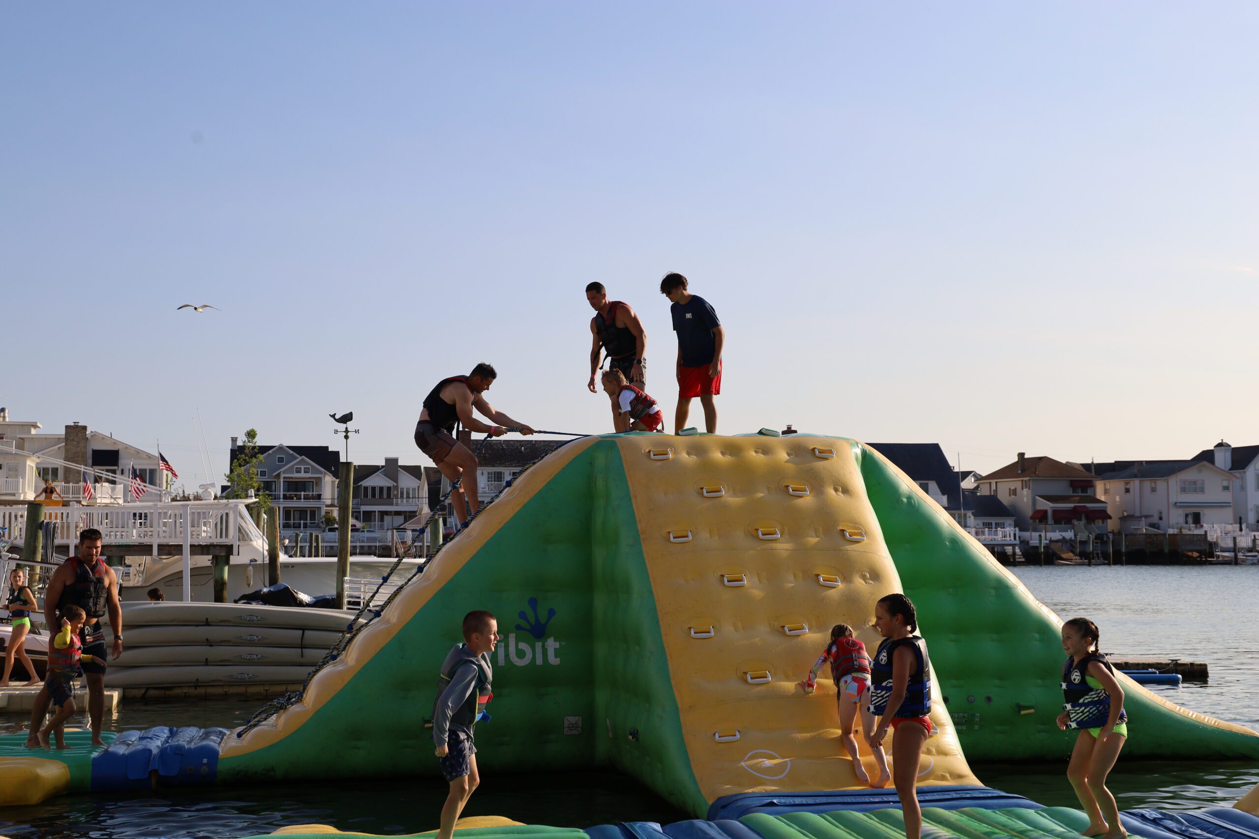 Island Water Sports lifeguard helping a young child up the rope of the inflatable climbing tower at the Stone Harbor aqua park while other kids play around the base