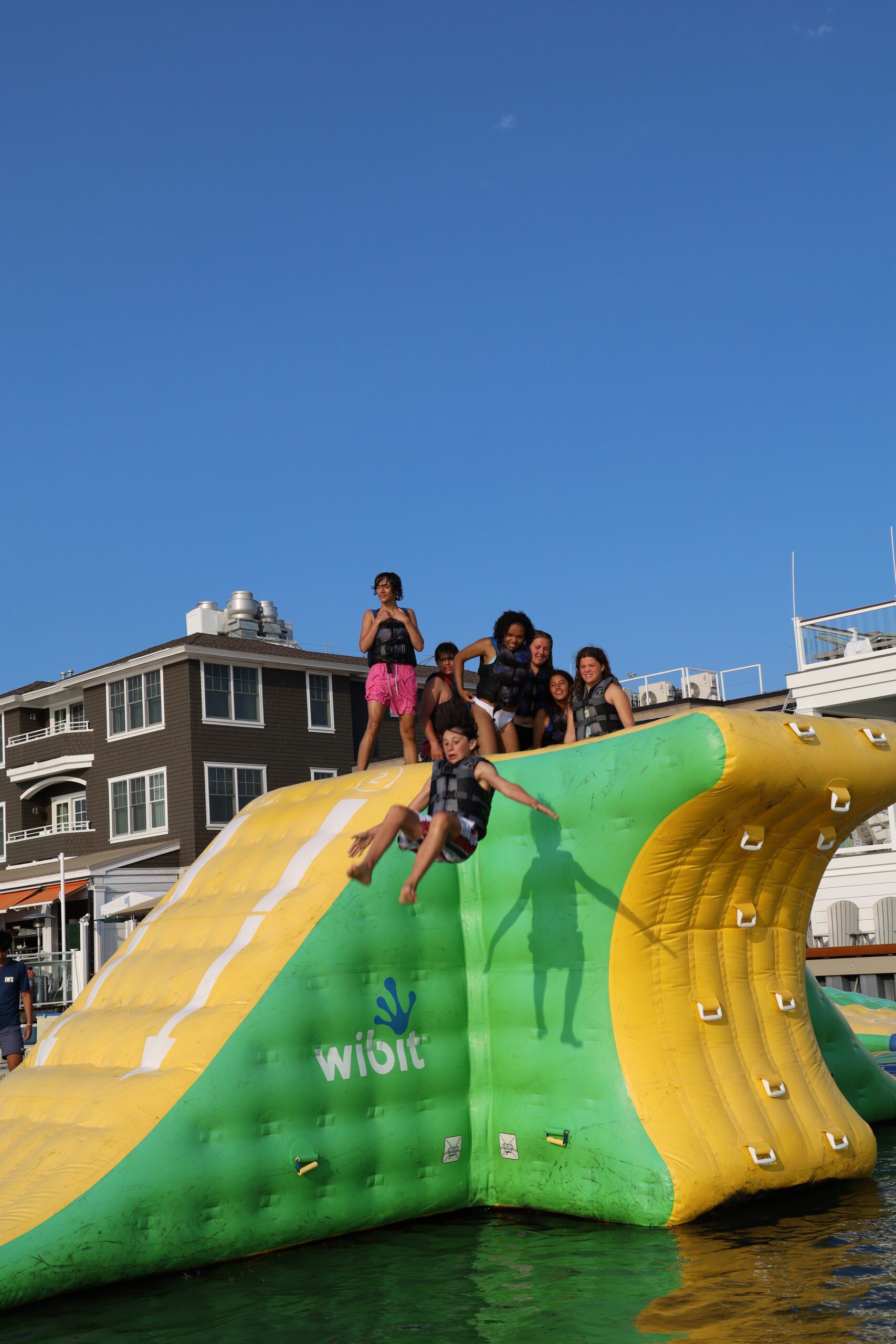 Boy in a life jacket sliding down a green and yellow inflatable slide at the Stone Harbor aqua park while a group of teens cheers from the top
