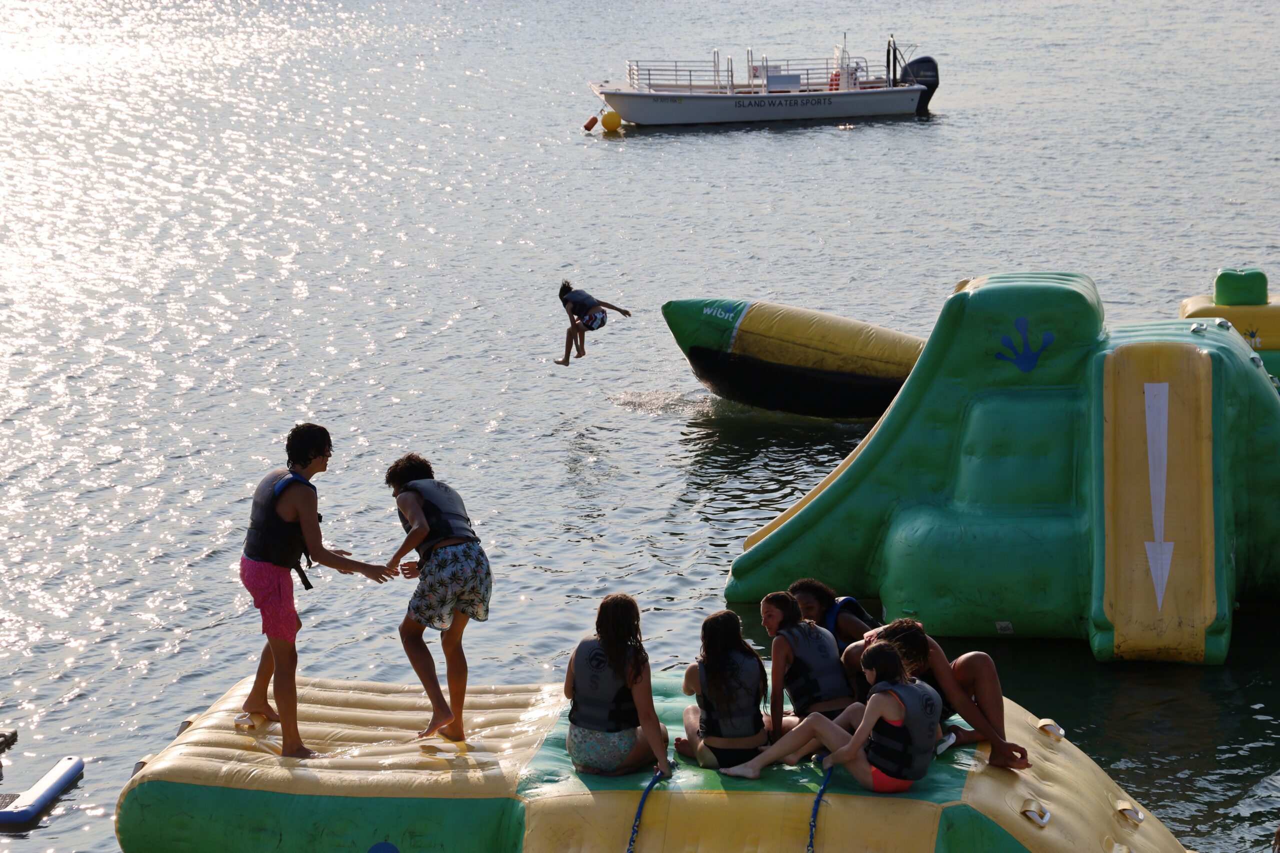 Kid leaping into the back bay at the Stone Harbor aqua park at sunset while a group of friends watches from the inflatable platform with the Island Water Sports boat in the background